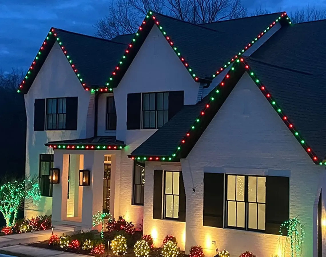 A white two-story home at dusk with permanent roofline lights alternating red and green for Christmas along every gable